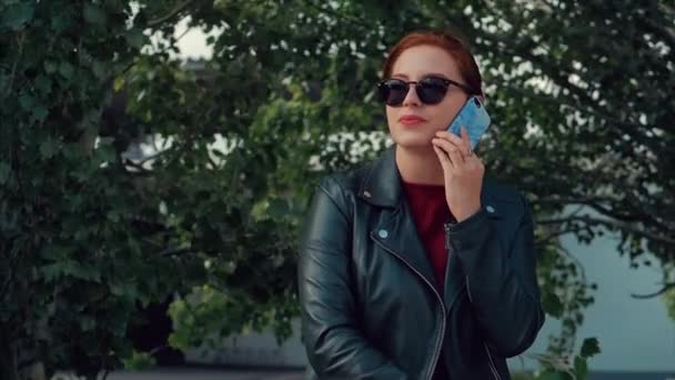 Молодая женщина в солнцезащитных очках разговаривает по мобильному телефону на улице, во время прогулки по городу . — стоковое видео