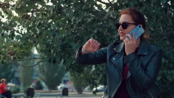 Jovem mulher falando no celular enquanto está sentada no parque em um banco, verifica as mensagens em seu relógio de pulso. Menina olha para horas quantas vezes enquanto fala ao telefone . — Vídeo de Stock