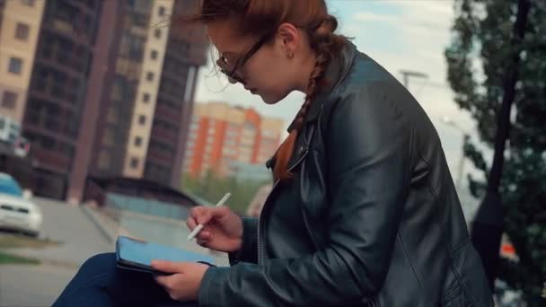 Mujer en gafas con el pelo largo y rojo en un buen estado de ánimo, emociones positivas, se basa en una tableta gráfica en el stree. Chica en el parque sentado en un banco, dibuja en una tableta gráfica . — Vídeos de Stock