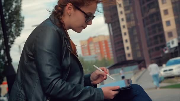 Женщина в очках с длинными рыжими волосами в хорошем настроении, положительные эмоции, рисует на графическом планшете на улице. Девушка в парке сидит на скамейке, рисует на графическом планшете . — стоковое видео