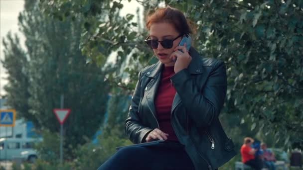 Mujer joven en gafas de sol hablando en el teléfono móvil mientras está sentado en el parque en un banco, registros de conversación en notas en una tableta. Chica hablando por teléfono . — Vídeos de Stock