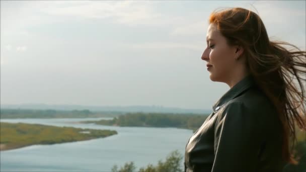 Portret van een kleine vrouw met rood haar staande hoog op een heuvel, meisje de achtergrond van de natuur, schilderachtig landschap met rivier in de verte, prachtige zonsondergang. wind speelt haar van een mooie vrouw. — Stockvideo