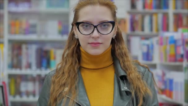 Retrato de una joven guapa sonriente joven con gafas mirando a la cámara en la biblioteca de la Universidad de la ciudad . — Vídeos de Stock