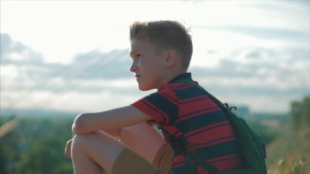 Porträt eines jungen kaukasischen Teenagers in einem roten Hemd mit einem Rucksack auf dem Rücken, bei Sonnenuntergang, auf einem hohen Hügel sitzend, den warmen Sommerurlaub genießend. — Stockvideo