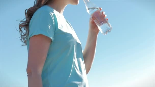Mujer de aspecto europeo al aire libre bebe agua de una botella contra un cielo azul claro. Estilo de vida saludable . — Vídeo de stock