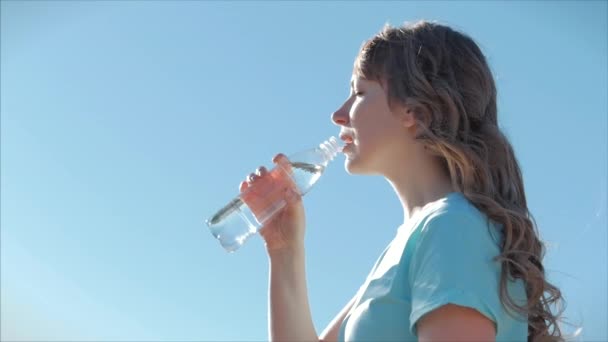 Femme d'apparence européenne en plein air boit de l'eau d'une bouteille contre un ciel bleu clair. Mode de vie sain . — Video