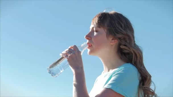 Mulher de aparência europeia ao ar livre bebe água de uma garrafa contra um céu azul claro. Estilo de vida saudável . — Vídeo de Stock