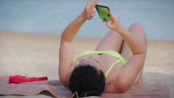 Europese vrouw ligt op het strand in een badpak, ligt te zonnebaden, ontspant, gebruikt telefoon, sms 't op een mobiele telefoon. Ontspanning concept, ontspannen op de zee, liggend op het zand. — Stockvideo