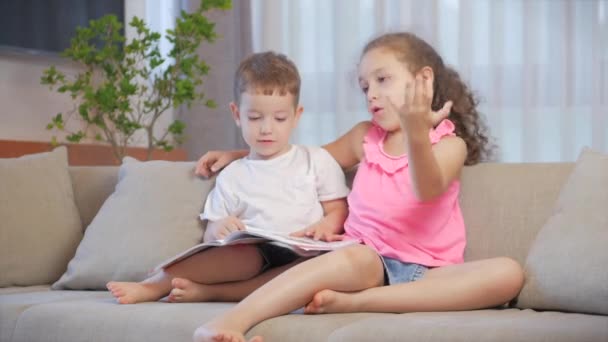 Lindos niños, hermana con un hermano menor, mirando una revista de libros diccionario de libros tableta leer cuentos de hadas, los niños están leyendo libros sentados en el sofá, la educación preescolar y escolar. — Vídeos de Stock