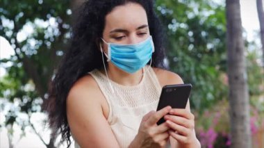 Koronavirüs salgınından maske takmış bir kadın parkta tek başına oturuyor, telefon kullanıyor, müzik kulaklığı kullanıyor, akıllı telefonundan mesaj yazıyor, küresel salgından kendini soyutlayan insanlar kavramını kullanıyor.