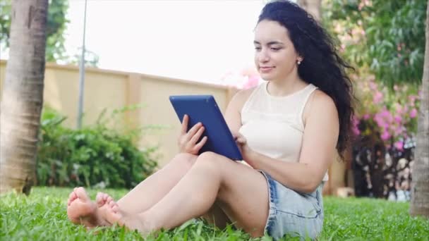 Europeiska kvinnan använder sig av en surfplatta eller telefon med en smartphone, blad eller sms med fingret, kommunicerar med hjälp av applikationen, kopplar av på gatan medan hon sitter på gräset i parken. — Stockvideo