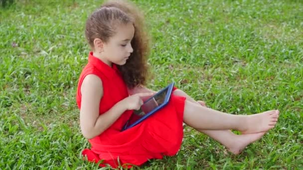 Kíváncsi aranyos lány óvodás gyermek egy digitális tablet technológia eszköz néz telefon ül az utcán a füvön a parkban. A gyerek a tabletben játszik.. — Stock videók