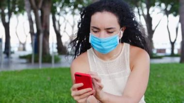 Koronavirüs salgınından maske takmış bir kadın parkta tek başına oturuyor, telefon kullanıyor, müzik kulaklığı kullanıyor, akıllı telefonundan mesaj yazıyor, küresel salgından kendini soyutlayan insanlar kavramını kullanıyor.