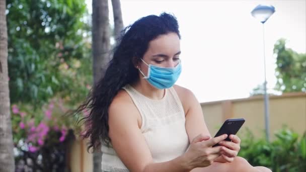 Frau mit Maske von der Coronavirus-Epidemie sitzt einsam im Park, hört per Kopfhörer Musik, tippt auf ihrem Smartphone SMS, entwirft ein Bild von einem Volk, das sich vor der globalen Epidemie selbst isoliert