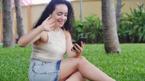 Mulher europeia faz uma chamada de vídeo on-line usando um smartphone via fones de ouvido conferência no telefone conversando usando uma webcam chamada de vídeo bate-papo na rua sentado na grama no parque . — Vídeo de Stock