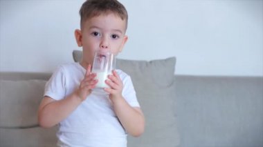 Evdeki kanepede oturup süt içen tatlı bebek. Yavaş çekim su içen küçük çocuk. Yakın plan. Komik küçük çocuk bir bardak su içiyor..