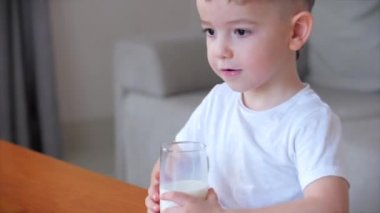 Evdeki kanepede oturup süt içen tatlı bebek. Yavaş çekim su içen küçük çocuk. Yakın plan. Komik küçük çocuk bir bardak su içiyor..