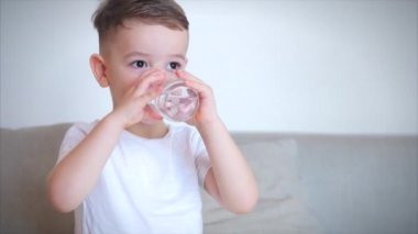 Evdeki kanepede oturup bir bardak su içen tatlı bir oğlan. Yavaş çekim su içen küçük çocuk. Yakın plan. Komik küçük çocuk bir bardak su içiyor..