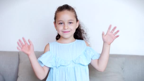 Retrato de uma menina engraçada sorrindo. Calma, a olhar para a câmara, a acenar as mãos. Olá como um blogueiro sentado em um sofá em casa, bebê bonito criança com um rosto bonito sentado na aula de polegar sofá — Vídeo de Stock