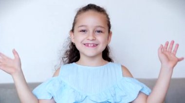 Gülümseyen komik küçük bir kızın portresi. Sakin ol, kameraya bak, el salla. Evdeki kanepede oturan bir blogcu olarak merhaba, şirin yüzlü şirin bir çocuk sofshaw başparmağında oturuyor.