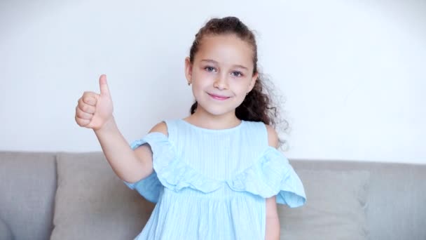 Divertido retrato niña sonriente niño mirando a la cámara sentarse en el sofá en casa, lindo niño con cara linda sentado en el sofá muestra pulgar hacia arriba clase. Concepto de una infancia feliz . — Vídeo de stock