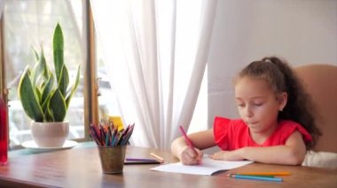 Mutlu küçük kız ya da 7-8 yaşlarındaki şirin ilkokul çocuğu evde oturup kâğıt kalem kullanarak resim yapıyor. Çocuk çizimleri, odaklanmış çocuklar ilköğretim konsepti.