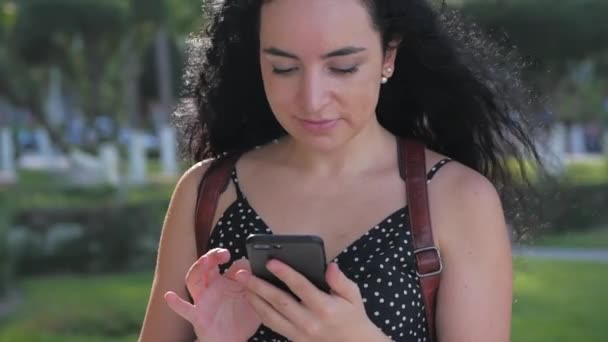 Young woman or girl is texting messages on the phone on a sunny day on a background of palm trees, greenery, trees on a city park. — Stock Video