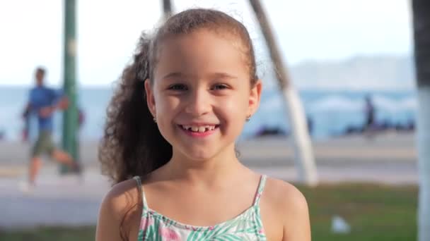 Kamera tebessümlerine bakan komik küçük bir kızın portresi, güzel bir gülümseme, güneşli bir yaz günü, anaokulunun şirin bebek yüzü, sokakta kameraya bakıyor.. — Stok video