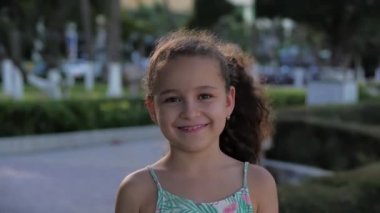 Kamera tebessümlerine bakan komik küçük bir kızın portresi, güzel bir gülümseme, güneşli bir yaz günü, anaokulunun şirin bebek yüzü, sokakta kameraya bakıyor..