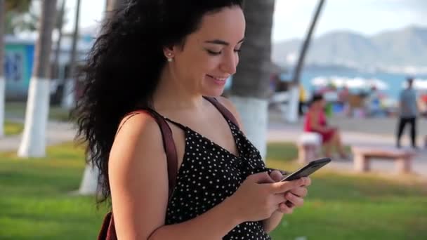 Девушка или девушка отправляет сообщения по телефону в солнечный день на фоне пальм, зелени, деревьев в городском парке. — стоковое видео