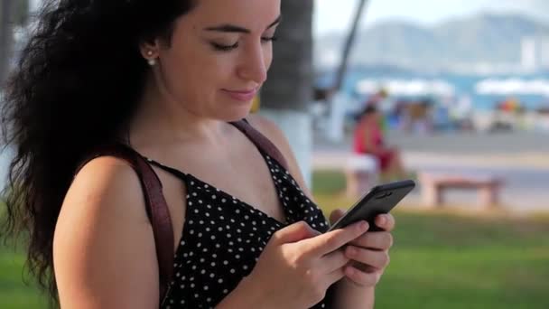 Mujer joven o niña está enviando mensajes de texto por teléfono en un día soleado sobre un fondo de palmeras, vegetación, árboles en un parque de la ciudad . — Vídeos de Stock