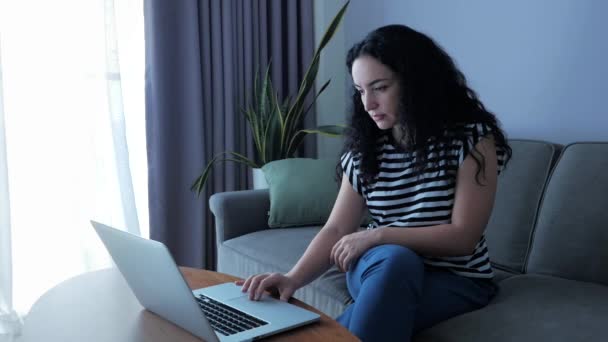 Giovane donna seria che lavora sul computer portatile, seduta sul divano a casa, la donna d'affari si siede a casa lavora a distanza sul computer portatile o smartphone controlla la posta per studiare il lavoro online a casa. — Video Stock