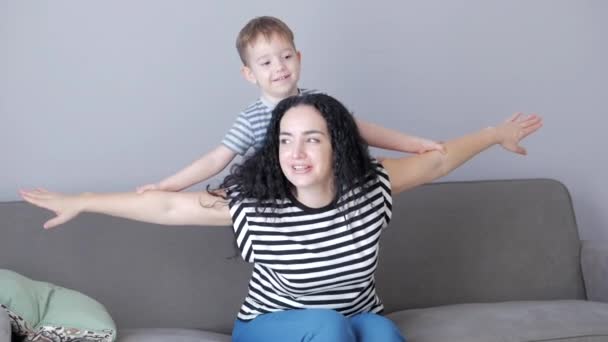 Happy healthy family young mom lifting cute little child son up playing plane on sofa at home, funny kid boy flying in mothers practice sport. — Stock Video