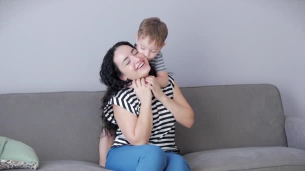 Gelukkig gezond gezin jong mam knuffels schattig klein kind zoon die kust moeder op de lippen of wang zitten op de bank thuis, grappig zoon jongen jongen liefde spelletjes moeder. — Stockvideo