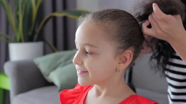 Mamá de una familia feliz, una amante europea en casa en el sofá haciendo peinado, peinando el pelo de su pequeña hija linda, el niño se regocija por la preocupación de las papas por ella . — Vídeo de stock