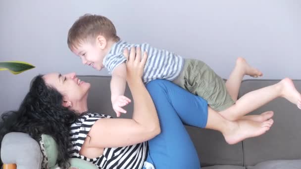 Feliz família saudável jovem mãe levantando bonito criança filho até jogando avião no sofá em casa, garoto engraçado voando em mães praticar esporte . — Vídeo de Stock