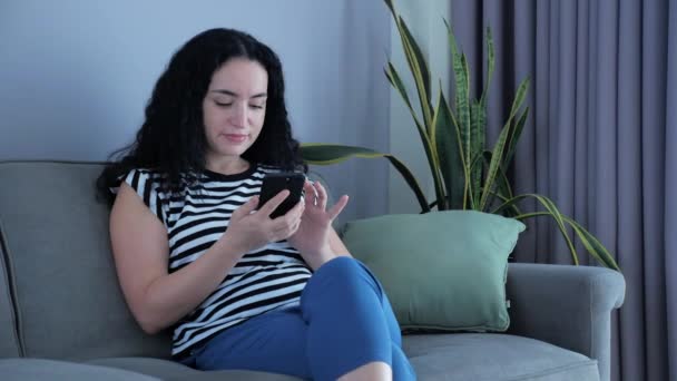 Mujer escribiendo en el teléfono sentado en el sofá en casa, mujer de negocios se sienta en los tipos de casa en cheques de teléfonos inteligentes correo para estudiar trabajo en línea en casa. — Vídeos de Stock