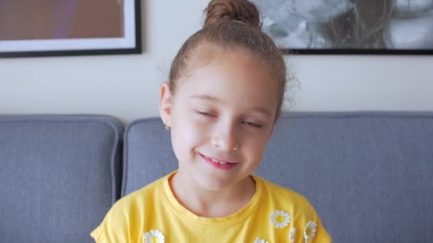 Portrét Legrační holčička usmívající se dítě dívající se do kamery sedí doma na gauči, roztomilé dítě s hezkou tváří sedí na pohovce. Koncept šťastného dětství. — Stock video