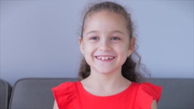 Portre Komik küçük kız kameraya bakıyor evde kanepede oturuyor, güzel yüzlü şirin çocuk kanepede oturuyor. Mutlu bir çocukluk kavramı.