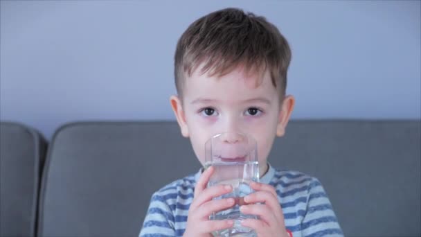Portrait Drôle petit enfant boit une tasse d'eau.Bébé garçon mignon buvant un verre d'eau assis sur le canapé à la maison. Petit garçon buvant de l'eau. Gros plan. — Video