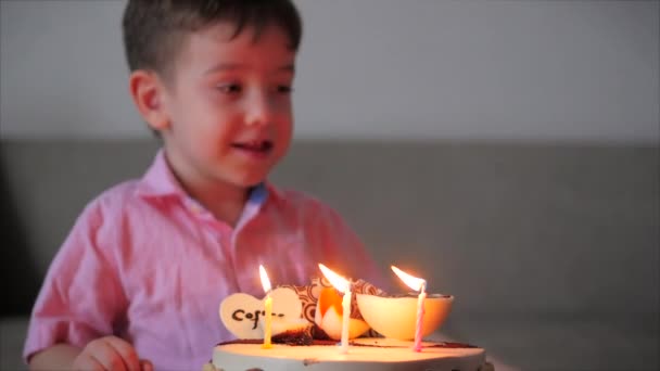 Щасливий маленький хлопчик з чотирьох сидить на дивані, дивлячись на палаючі свічки на торт на день народження, робить побажання на день народження, святкує день народження зі своєю сім'єю, і вибухає свічки на торт . — стокове відео