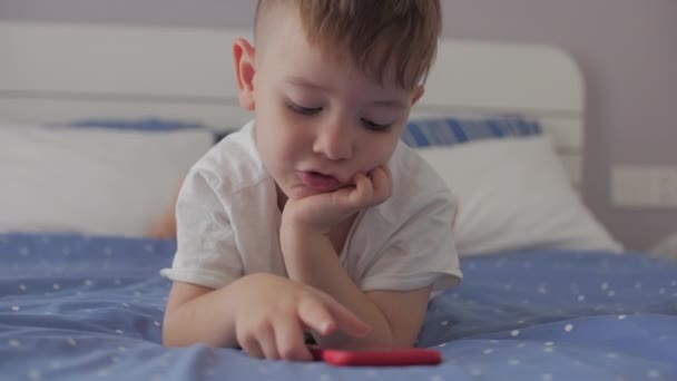 Lindo niño preescolar niño usando un teléfono o tableta digital dispositivo de tecnología se ve teléfono, juega en el teléfono sentado resto tumbado en el sofá solo.Children tech addiction concept play game in home. — Vídeo de stock