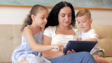 Mutlu aile, anne ve tatlı çocuklar tablet ya da dizüstü bilgisayar izliyor, anne evde çocuklarla oynuyor, tablet kullanıyor, sarılıyor, kanepede oturuyor, gülüyor, eğleniyor, aile hayatının tadını çıkarıyor..