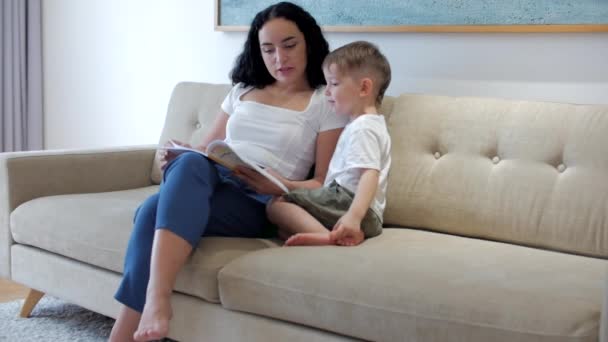 快乐的家庭，妈妈和可爱的学龄前男孩，儿子和妈妈一起读书，坐在沙发上，妈妈读小说，享受家庭时光. — 图库视频影像