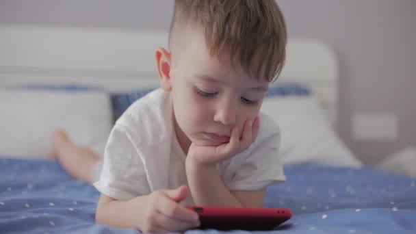 Söt liten pojke förskola barn med hjälp av en telefon eller digital surfplatta teknik ser telefon, spelar i telefonen sittande vila ligger på soffan ensam.Barn tech missbruk koncept spela spel hemma. — Stockvideo