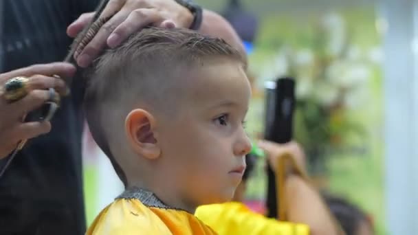 Kinderfrisur im Salon zur Zeit der Coronavirus-Epidemie. Kleiner Junge bekommt einen Haarschnitt. Kinderhaarstyling. Stylistin in Schutzmaske von covid-19 sorgt für stylische Frisur. — Stockvideo