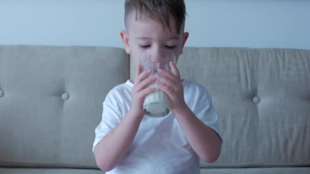 Bonito menino bebendo um copo de leite sentado no sofá em casa. Rapazinho de câmara lenta a beber água. Close-up. Retrato Engraçado Criança está bebendo um copo de água. — Vídeo de Stock