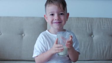 Evdeki kanepede oturup süt içen tatlı bebek. Yavaş çekim su içen küçük çocuk. Yakın plan. Komik küçük çocuk bir bardak su içiyor..