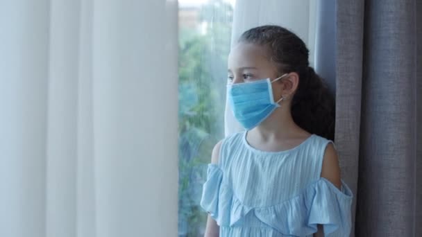 Uma criança pequena ou uma menina em uma máscara médica protetora olha para fora da janela por trás de uma cortina, com um rosto triste e decepcionado. Uma rapariga com uma máscara médica. Pandemia, covid - 19 — Vídeo de Stock