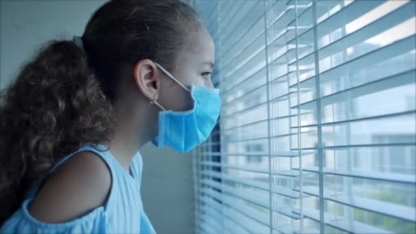 Portrét roztomilé malé dítě nebo holčička v ochranné lékařské masce se dívá z okna, se smutnou zklamanou tváří. Dívka v lékařské masce. Pandemie, covid - 19 — Stock video